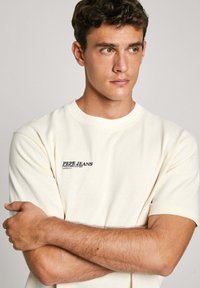 Camiseta de algodón color crema con mangas cortas, que presenta un logo impreso en negro en el área del pecho izquierdo. Textura suave y corte relajado.