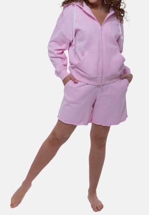 Vrouw draagt een lichtroze hoodie met ritssluiting en bijpassende korte broek, staat blootsvoets met handen in de zakken en één been opzij geheven.