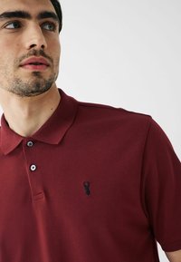 Bordo polo majica sa teksturiranim materijalom, s crnim izvezenim logom i tri dugmeta na ovratniku. Kratki rukavi.