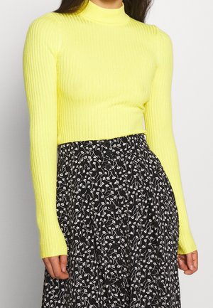 Femme portant un haut à manches longues côtelé jaune vif, associé à une jupe taille haute noire ornée de petites fleurs blanches.