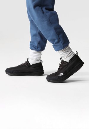 Personne portant des baskets noires The North Face, des chaussettes blanches et un pantalon bleu, avançant sur un fond clair uni.