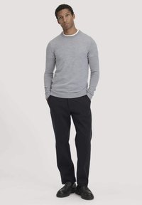 Hellgrauer Langarmpullover mit Rundhalsausschnitt, kombiniert mit schwarzen Hosen und schwarzen Schuhen. Der Pullover verfügt über gerippte Bündchen und einen gerippten Saum.