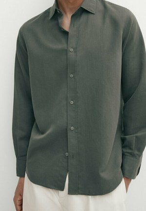 Homme portant une chemise verte foncée à manches longues boutonnée, entrée de manière lâche dans un pantalon blanc, debout devant un fond uni.