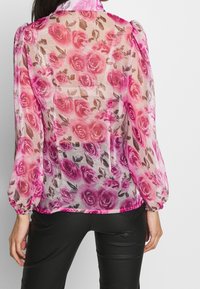 Gennemsigtig pink bluse med blomstret rosemønster, puffede ærmer og en let buet kant. Fremstillet af let, semitransparent stof.