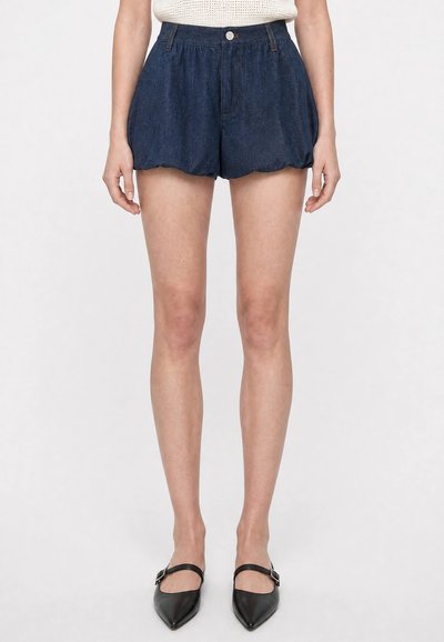 Femme portant un short en denim foncé taille haute avec un bouton et des poches avant, associé à des chaussures Mary Jane noires à bout pointu.