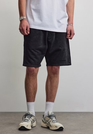 Homme portant un short noir, des chaussettes blanches montantes et des chaussures de course beiges, debout devant un mur et un sol unis.