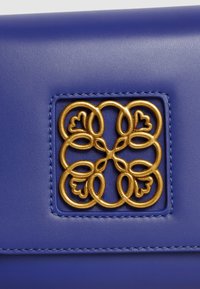 Borsa in pelle blu reale con un logo in metallo dorato testurizzato caratterizzato da design circolari intrecciati e cuciture decorative.