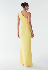 Gul, en-skulder kjole med en tætsiddende silhuet, tekstureret stof og lang længde, der har en drapering bagpå og minimale skodetaljer.