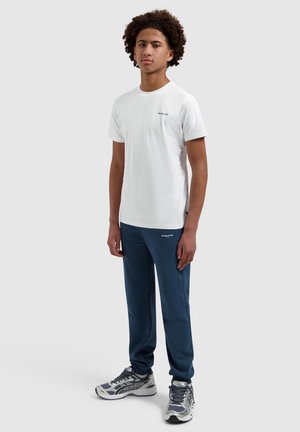 Jonge man staand, met een wit T-shirt met korte mouwen, blauwe joggingbroek en grijze sportschoenen op een lichtgrijze achtergrond.