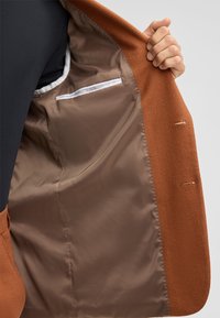 Brauner Jackeninnenraum, glatter satinähnlicher Futter, linker Seitentasche mit weißem Besatz, traditioneller Reversstichdetails.
