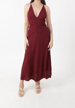 Femme portant une robe midi bordeaux sans manches à décolleté profond en V, avec des sandales beige à bride autour de la cheville, debout sur fond blanc.