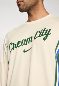 Výkonnostný dres krémovej farby s nápisom „Cream City“ v zelenej farbe, s logom Nike Swoosh a modro-zelenými akcentmi po stranách.