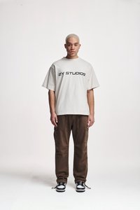 T-shirt oversized crema con la scritta "2Y STUDIOS" in lettere nere, abbinata a pantaloni cargo marroni e sneakers bianco e nero. Sfondo semplice.