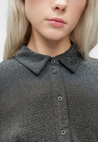 Artimų planų nuotrauka, kurioje matyti asmuo, vilkintis juodai baltą taškuotą marškinį su plačia apykakle ir šviesiai blondiniais plaukais.