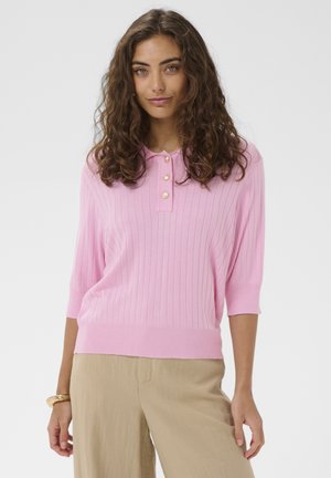 Femeie cu păr creț purtând un pulover polo tricotat roz cu nasturi aurii și pantaloni bej, stând pe un fundal simplu.