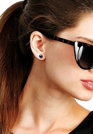 Jeune femme aux cheveux bruns attachés en queue de cheval, portant des boucles d'oreilles clous noires et de grandes lunettes de soleil écaille de tortue, tournée vers la droite.