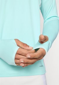 Šviesiai mėlyna ilgomis rankovėmis marškinėliai su nykščio angomis, lygiu paviršiumi ir subtiliais siūlais. Rodomos rankos, kurios reguliuoja rankovę.