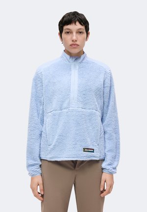 Person med kort mørkt hår iført lyseblå tekstureret pullover med lynlås foran og beige bukser, står mod en ensfarvet baggrund.