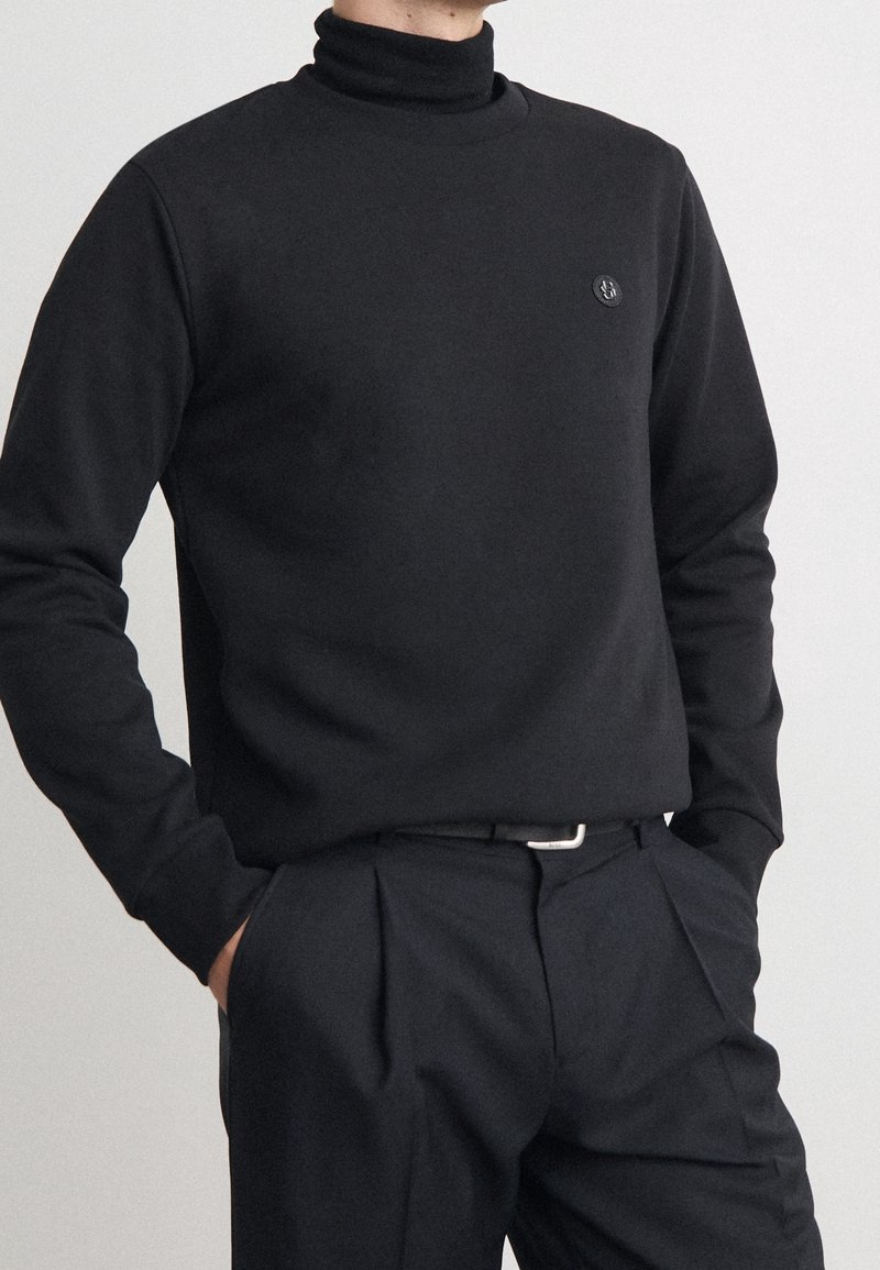 Černý rolákový svetr z měkké látky s úzkým střihem, s malým emblémem na hrudi. Kombinováno s černými kalhotami s plisováním.