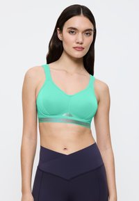 Ung kvinde med langt mørkt hår iført en turkis sports-bh og mørkeblå træningsleggings med høj talje mod en ensfarvet hvid baggrund.