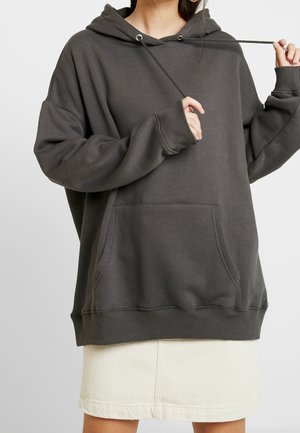 Kobieta nosząca oversize ciemnoszarą bluzę z kapturem z przednią kieszenią i białą spódnicę, trzymając rękami jeden sznurek od bluzy.