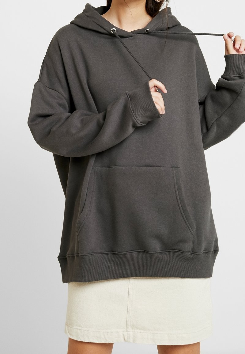 Femme portant un sweat à capuche gris foncé oversize avec poche avant et jupe blanche, tenant un cordon de capuche avec ses mains.