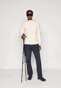 Kreemikleit fliisjakk, millel on kõrge krae, koos tumedate cargo-pükstega. Modell hoiab musta matkasau, kannab tumedaid päikeseprille ja matkasaapaid.