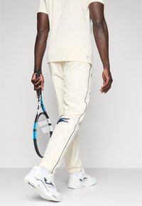 Pantaloni jogger crema con accenti blu navy e logo a coccodrillo, abbinati a scarpe da ginnastica bianche e una racchetta da tennis nera.