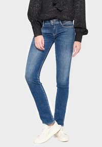Jeans skinny en denim bleu moyen, présentant un design classique à cinq poches, des passants de ceinture et des coutures contrastées, associés à des baskets blanches.