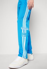 Blå Adidas träningsbyxor med vita ränder och knappar, bärs med vita sneakers av en stående person.