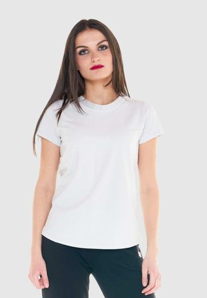 Giovane donna con lunghi capelli castani che indossa una camicia bianca a maniche corte e pantaloni neri su uno sfondo chiaro e uniforme.