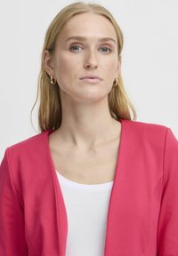 Valkoinen topin päällä oleva kirkkaan vaaleanpunainen bleiseri V-pääntie-muotoilulla, joka on valmistettu sileästä kankaasta. Minimalistiset linjat ja siistit reunat.