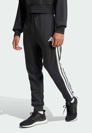 Homme portant un pantalon de survêtement Adidas noir avec des bandes blanches et des baskets noires, debout avec un pied légèrement en avant.