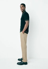 Young man standing sideways wearing a black short-sleeve polo, beige pants, black shoes, with hands in pockets against a white background.