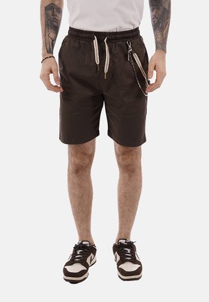 Uomo con pantaloncini neri con coulisse e accessorio a catena, sneakers bianche e tatuaggi visibili sulle braccia, in piedi contro uno sfondo bianco.