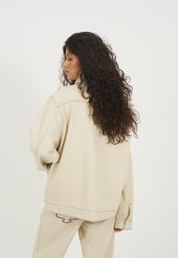 Beige, oversized dzseki látható varrási részletekkel, esett vállakkal és lazább szabással, hátulról, sima fehér háttérrel bemutatva.