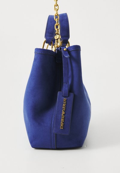 Mėlyna zomšinė rankinė su auksinėmis grandinėlėmis ir auksiniu „EMPORIO ARMANI“ ženkleliu, kabantčiu ant paprasto balto fono.