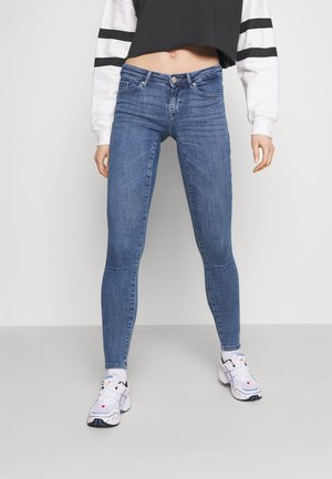 Frau trägt blaue Skinny-Jeans, schwarzes und weißes kurz geschnittenes Langarmshirt, weiße Socken und weiße Laufschuhe vor schlichtem grauem Hintergrund.