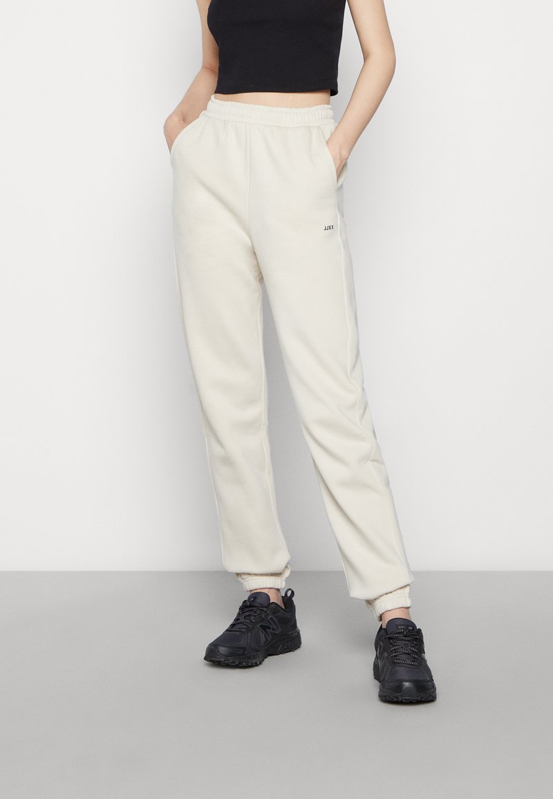 Cremfarbene Jogginghose mit elastischem Bund und Bündchen, ausgestattet mit Seitentaschen und einem kleinen schwarzen Logo. Tragen mit schwarzen Sneakers.