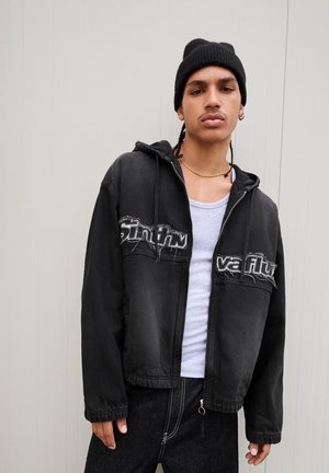 Black cropped jacket with a hood, featuring distressed lettering on the front. Worn over a gray t-shirt and paired with dark jeans.