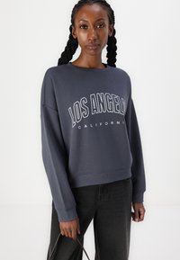Felpa grigia scura con testo "LOS ANGELES CALIFORNIA" in bianco. Presenta un collo a giro e maniche lunghe, abbinata a jeans neri.