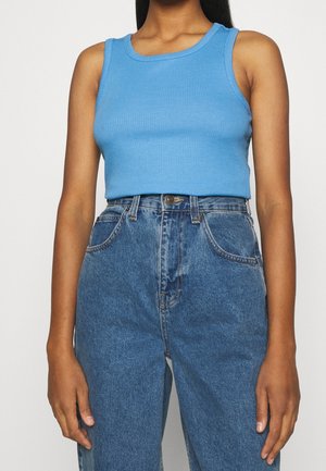 Vrouw draagt een mouwloze, geribbelde blauwe crop top en hooggetailleerde, losse blauwe spijkerbroek tegen een effen achtergrond.