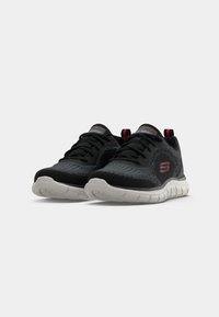 Black athletic shoes with a mesh upper, white textured sole, red accents, and a logo on the side. Laces and pull tab included.
