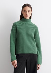 Grüner, gerippter Strickpullover mit hohem Kragen und langen Ärmeln, in entspannter Passform und mit seitlichen Schlitzen, kombiniert mit schwarzen Hosen.