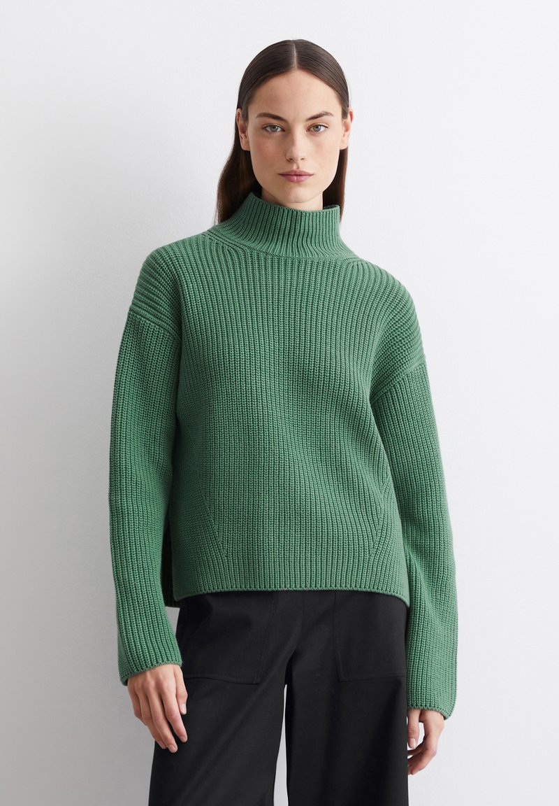 Grüner, gerippter Strickpullover mit hohem Kragen und langen Ärmeln, in entspannter Passform und mit seitlichen Schlitzen, kombiniert mit schwarzen Hosen.