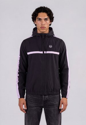 Jeune homme aux cheveux noirs et bouclés, portant une veste noire à zip avec des bandes roses et un jean foncé, debout devant un fond uni.