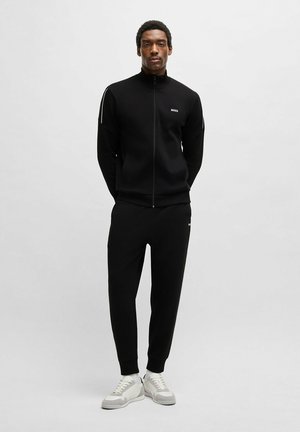 Zwart sportief trainingspak met een ritsjack, geribbelde manchetten en bijpassende broeken. Bevat een logo op de borst en zijstrepen. Witte sneakers.