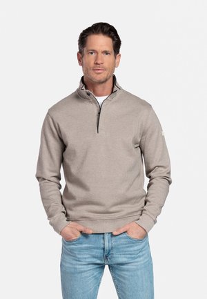 Man met een beige trui met kwart-ritssluiting en lichtblauwe spijkerbroek staat met zijn handen in zijn zakken tegen een witte achtergrond.