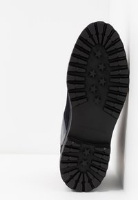 Semelle d'une botte noire avec des crampons profonds et des motifs en forme d'étoile, inclinée contre un coin blanc en arrière-plan.