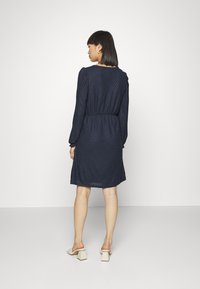 Vestido azul-marinho com mangas longas, tecido texturizado, cintura ajustada, comprimento até ao joelho, usado com mules de tacão quadrado de cor clara.